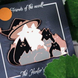 May include: Rose gold enamel pin featuring a white cat wearing a witch hat with a group of black and gray cats surrounding it. The pin says "Friends of the occult" and "The Hunter's" at the top and bottom.