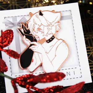 May include: A white enamel pin featuring a black and white illustration of a person with horns and a bloody mouth. The pin is displayed on a white background with a silver ribbon.