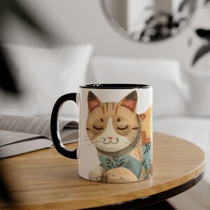 Puede incluir: Una taza de cerámica blanca con asa y borde negros. La taza presenta una ilustración a la acuarela de un gato relajado con los ojos cerrados, que lleva una manta estampada. El gato es de color canela y blanco con rayas marrones. La taza está sobre una mesa de madera.