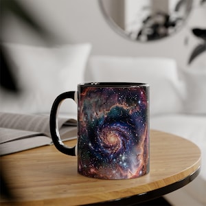 Könnte beinhalten: Keramikbecher mit einem Galaxiemuster in Lila, Blau und Orange. Der Becher hat einen schwarzen Innenraum und Henkel. Das Design zeigt einen wirbelnden Nebel.