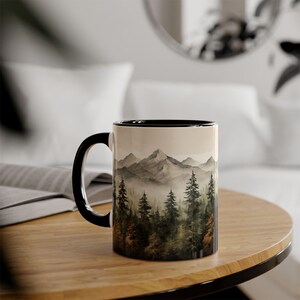 May include: A ceramic coffee mug with a black handle and rim. The mug features a scenic landscape of mountains and trees in shades of green, brown, and gray. The design wraps around the mug, creating a cohesive image. The mug sits on a wooden table.