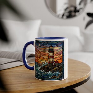 May include: White ceramic mug with a blue handle and rim. The mug features a colourful stained-glass style design of a lighthouse on a rocky coast with a sunset background. The lighthouse is red and white.