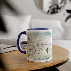 Op de afbeelding: Witte keramische mok met een blauw handvat en rand. De mok heeft een aquarelontwerp van paardenbloemzaden en de woorden "...just breathe" in zwart schrift. De mok staat op een houten tafel.