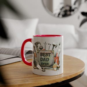 May include: A white ceramic mug with a red interior and handle. The mug features a design of tools and the text "BEST DAD EVER" on a metal sign. The mug is sitting on a wooden table.