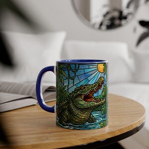 May include: A ceramic mug with a blue handle and rim. The mug features a colourful illustration of an alligator in a swamp scene with a blue sky and sun. The design has a stained glass effect.