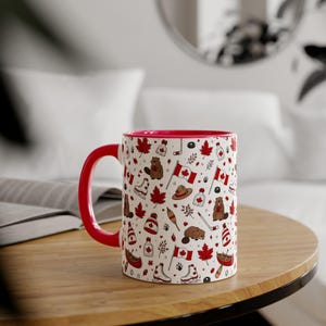 Può includere: Una tazza di ceramica bianca con manico e interno rossi. La tazza è decorata con un motivo di simboli canadesi, tra cui foglie d'acero, bandiere, castori e mazze da hockey. La tazza è su un tavolo di legno.
