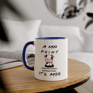 Peut inclure: Mug en céramique blanche avec une anse et un bord bleus. Le mug présente le texte "A MOO POINT" avec une illustration de vache de dessin animé. En dessous, le texte "It's like a cow's opinion: It just doesn't matter. IT'S MOO."