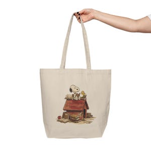 May include: Beige tote bag featuring a Snoopy and Woodstock graphic. The illustration shows Snoopy and Woodstock sitting on top of Snoopy's doghouse, surrounded by books. The bag has two handles and is held up by a person.