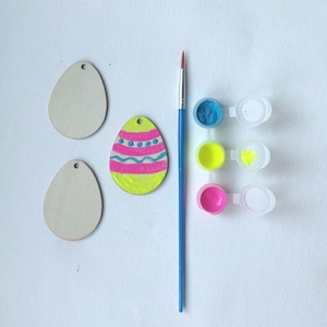 May include: An Easter egg painting kit with three wooden egg shapes, one painted egg, three paint pots in blue, yellow, and pink, and a paintbrush. The painted egg has pink and blue stripes and dots on a yellow base.