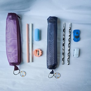 Puede incluir: Un estuche de malla morada con cremallera, un lápiz color melocotón, un lápiz plateado, una goma de borrar azul, un sacapuntas con forma de flor, un estuche de malla azul marino, dos lápices con diseños de balón de fútbol, un coche de juguete azul y un coche de juguete blanco.