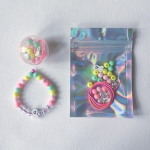 Op de afbeelding: Een pastelkleurige kralenarmband met een witte spookbedel, een doorzichtige plastic bal met kralen en een spookbedel, en een holografische zak met ritssluiting met kralen en elastisch touw. De armband is gemaakt van roze, gele en groene kralen.