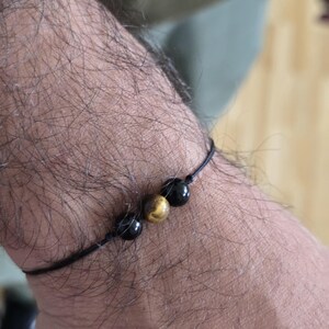 Puede incluir: Un brazalete de cuerda negra con tres cuentas: dos negras y una de ojo de tigre.