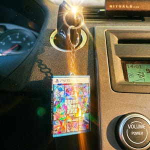 Può includere: Un portachiavi rettangolare che presenta una replica in miniatura della custodia del videogioco Grand Theft Auto V per PS5. Il portachiavi ha un anello e una catena in metallo argentato, e la copertina mostra il titolo con un design a collage colorato.