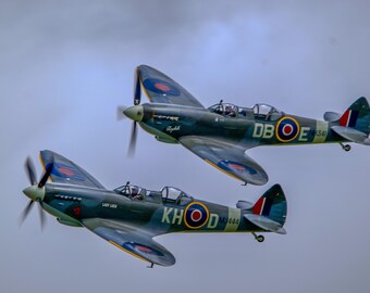 May include: Two vintage green and blue fighter planes flying in formation. The plane in the foreground has the text "KHOD" and "M4444" on the side. The plane in the background has the text "DBOE" and "E 1341" on the side.