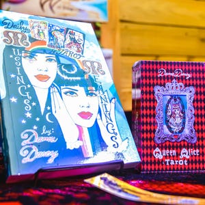 May include: A tarot card set and book are displayed on a red surface. The book cover features a blue and white illustration of a woman's face. The tarot card box is red with a black diamond pattern and the text "Queen Alice Tarot".