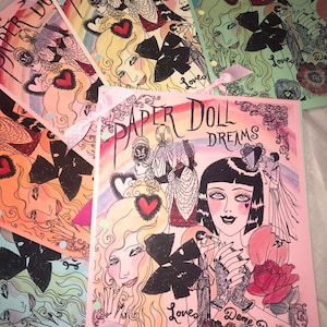 May include: A pink paper doll book with the title "Paper Doll Dreams" in black and white lettering. The book features illustrations of women with colorful clothing and flowers. The book has a pink ribbon tied around it.