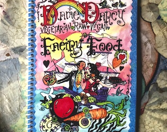 Könnte beinhalten: Ein spiralgebundenes Notizbuch mit blauem Glitzerrahmen und dem Titel "Dame Darcy Fairy Food". Das Cover zeigt eine farbenfrohe Illustration mit einem Regenbogen, einem Paar und verschiedenen Lebensmitteln. Die Worte "Vegetarian Raw Vegan" sind ebenfalls sichtbar.