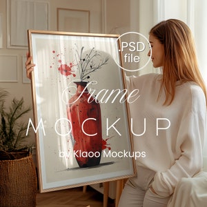 Op de afbeelding: Een vrouw houdt een ingelijst afdruk van een aquarel van een rode vaas met zwarte takken en rode spatten. De lijst is van hout met een lichtbruine afwerking. De tekst "Frame Mockup by Klaoo Mockups" is afgedrukt op de afdruk.