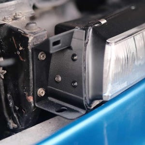 May include: Close-up of a car headlight assembly. The headlight is black with a clear lens, mounted on a blue car body. The assembly includes a black bracket secured with screws.