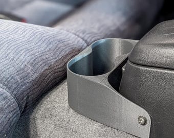 May include: A gray, 3D-printed car cup holder with two circular openings. The cup holder is positioned next to a black textured console and a gray fabric seat. The design is simple and functional, likely for in-car use.