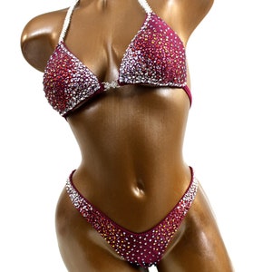 May include: Burgundy bikini with crystal embellishments. The top has a halter style with a silver clasp in the front. The bottom has a V-shaped cut.