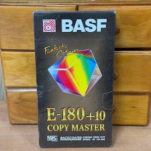 May include: A vintage BASF E-180+10 Copy Master VHS tape. The black box features the BASF logo, the words "Fantastic Colours", and a colorful geometric design. The tape is labeled "Backcoated Chrome Video Tape" and "Stereo SP: 190 MIN".