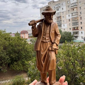 May include: A brown wooden figurine of a bearded person wearing a hat, a jacket, a vest, and trousers. The figure stands on a base and carries a long cylindrical object over the right shoulder, with the left hand resting on the hip area.