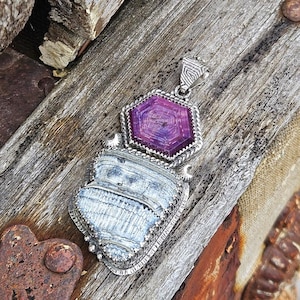Può includere: Un ciondolo in argento con una gemma esagonale viola incastonata sopra una pietra grigia testurizzata a forma di ventaglio. La montatura in metallo presenta un bordo decorativo a corda e una contromaglia triangolare con linee incise.