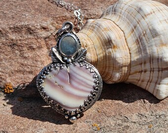 Puede incluir: Un collar de plata con un colgante en forma de corazón con una concha rosa y blanca y una piedra azul iridiscente. El collar está sobre una superficie marrón texturizada junto a una concha.
