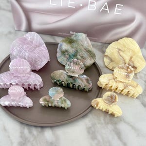 以下が含まれることがあります： ピンク、グリーン、イエローなど、さまざまな色の貝殻型のヘアクリップのコレクション。 クリップは茶色のプレートに配置され、背景には「LIE. BAE」と書かれたお揃いのバッグがあります。