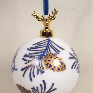 Op de afbeelding: Witte kerstbal met een blauw en goud dennenappel- en takontwerp. De ornament heeft een gouden hertenkop-opzetstuk en een blauw lint om op te hangen. Een feestelijke vakantieversiering.