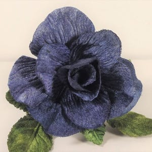 Könnte beinhalten: Eine dunkelblaue Stoffblume mit einer strukturierten, zerknitterten Optik auf den Blütenblättern. Die Blume zeigt eine zentrale Blüte mit mehreren geschichteten Blütenblättern und mehreren grünen, strukturierten Blättern am Stielansatz.