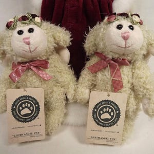 Puede incluir: Dos figuras de cordero de peluche color crema, cada una con una corona floral y un lazo de cinta rosa. Los corderos tienen ojos negros y narices rosas. Cada cordero tiene una etiqueta que dice "Lilith Angel Ewe".
