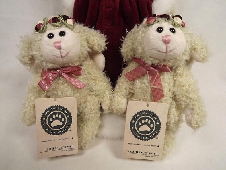 Puede incluir: Dos figuras de cordero de peluche color crema, cada una con una corona floral y un lazo de cinta rosa. Los corderos tienen ojos negros y narices rosas. Cada cordero tiene una etiqueta que dice "Lilith Angel Ewe".