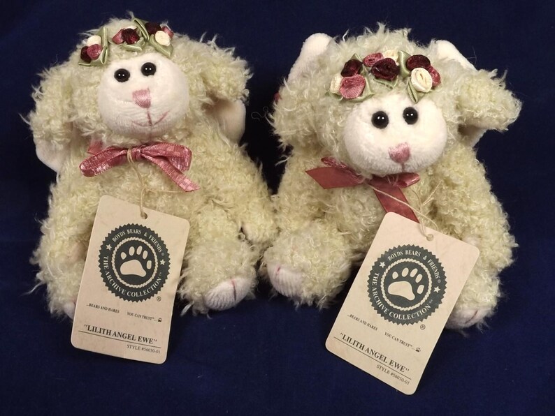 Puede incluir: Dos ovejas de peluche color crema, cada una con una corona floral y un lazo rosa. Los juguetes tienen ojos negros y narices rosas. Cada uno tiene una etiqueta que dice "Lilith Angel Ewe".