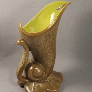 May include: A vintage ceramic vase with a distinctive design. The vase has a brown, textured exterior and a bright green interior. The base is leaf-shaped, and a curved accent enhances the artistic look. A decorative item for home decor.