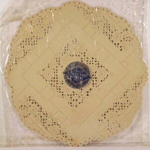May include: A white paper doily with a blue and white label that reads "DECORATED DOILIES" in a circle. The doily has a delicate lace pattern.