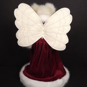 Puede incluir: Figura decorativa de &aacute;ngel con alas acolchadas de color crema y un vestido de terciopelo burdeos. El vestido est&aacute; adornado con piel sint&eacute;tica blanca. La cabeza del &aacute;ngel est&aacute; adornada con plumas blancas, creando una decoraci&oacute;n festiva.