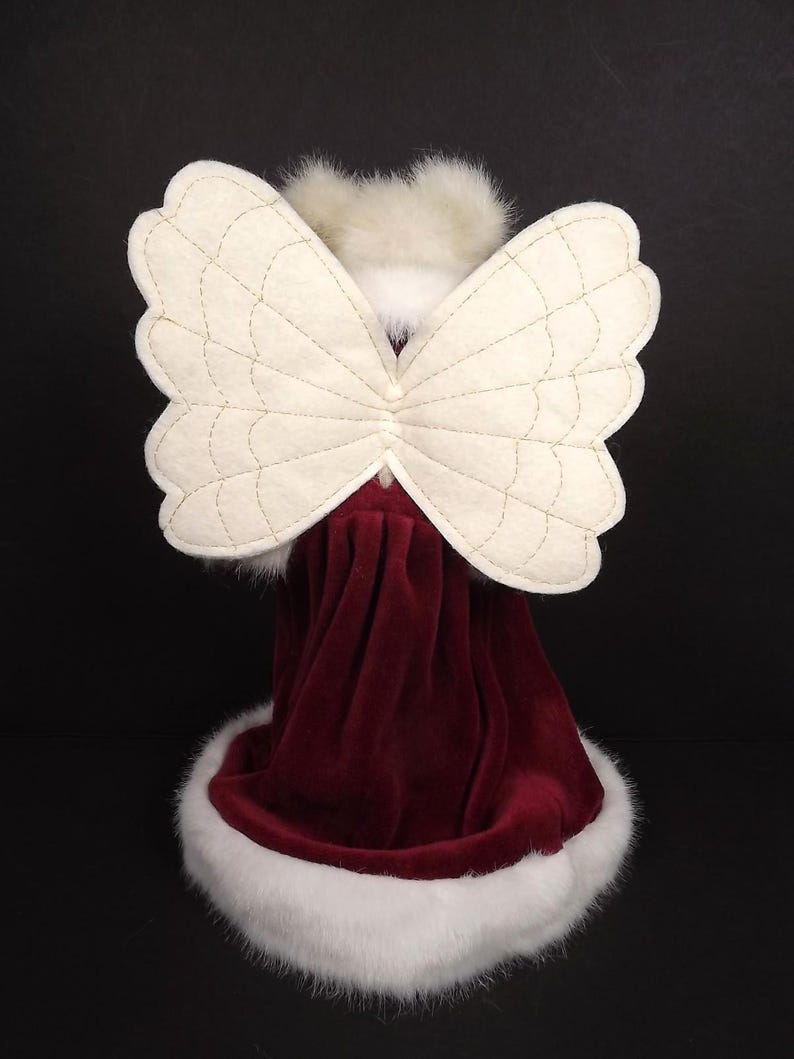 Puede incluir: Figura decorativa de &aacute;ngel con alas acolchadas de color crema y un vestido de terciopelo burdeos. El vestido est&aacute; adornado con piel sint&eacute;tica blanca. La cabeza del &aacute;ngel est&aacute; adornada con plumas blancas, creando una decoraci&oacute;n festiva.