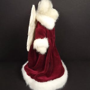 Puede incluir: Figura decorativa de &aacute;ngel con una t&uacute;nica de terciopelo burdeos con ribete de piel sint&eacute;tica blanca. El &aacute;ngel tiene un halo y alas de plumas blancas. Decoraci&oacute;n festiva navide&ntilde;a.