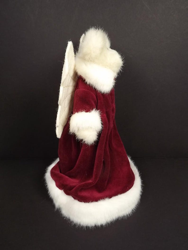 Puede incluir: Figura decorativa de &aacute;ngel con una t&uacute;nica de terciopelo burdeos con ribete de piel sint&eacute;tica blanca. El &aacute;ngel tiene un halo y alas de plumas blancas. Decoraci&oacute;n festiva navide&ntilde;a.