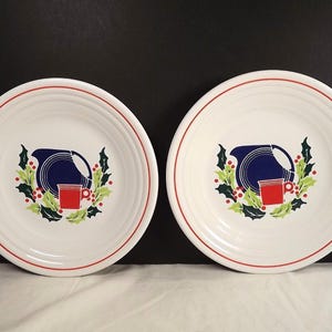 Puede incluir: Dos platos de cerámica blanca con borde rojo y un diseño central que presenta una jarra azul, una taza roja y hojas de acebo. Los platos están sobre un fondo oscuro.