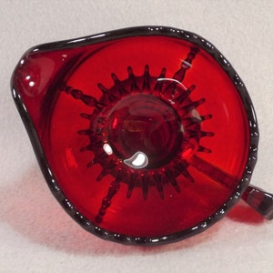 Puede incluir: Un exprimidor de c&iacute;tricos de vidrio rojo con un dise&ntilde;o de estrella en la parte inferior. El exprimidor tiene un asa y un pico.