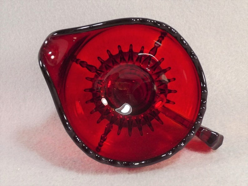 Puede incluir: Un exprimidor de c&iacute;tricos de vidrio rojo con un dise&ntilde;o de estrella en la parte inferior. El exprimidor tiene un asa y un pico.