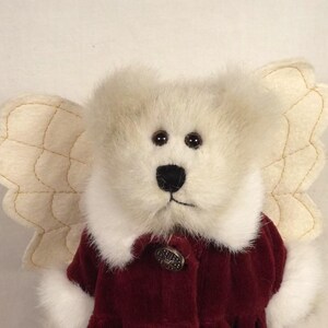 Puede incluir: Un oso de peluche &aacute;ngel con pelaje color crema y alas cosidas de color beige. El oso lleva un abrigo de terciopelo burdeos con cuello blanco y un bot&oacute;n decorativo. El oso tiene ojos marrones oscuros y una nariz negra.