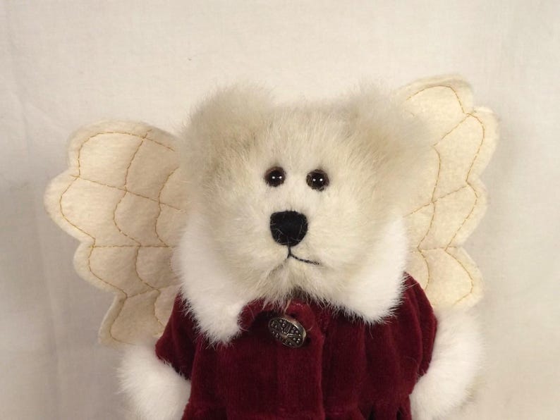 Puede incluir: Un oso de peluche &aacute;ngel con pelaje color crema y alas cosidas de color beige. El oso lleva un abrigo de terciopelo burdeos con cuello blanco y un bot&oacute;n decorativo. El oso tiene ojos marrones oscuros y una nariz negra.