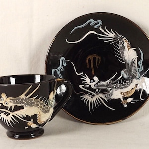 May include: A black teacup and saucer set with a white dragon design. The dragon is painted in white, gold, and blue on a black background.