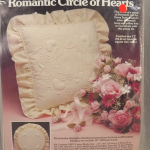 Peut inclure: Un kit de coussin blanc avec un motif de cercle de cœurs. Le kit comprend du coton mousseline 100%, du ruban biais, de la dentelle, du fil à broder, de la laine, du rembourrage et des instructions. La taille finale du coussin est de 43,2 cm de diamètre ou de côté.