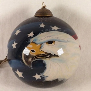 May include: A round Christmas ornament featuring a painted bald eagle and American flag design. The ornament has a dark blue background with white stars and a brass-colored cap. The eagle is painted in white, brown, and blue.