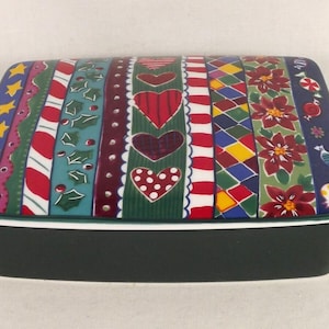 Puede incluir: Una caja rectangular de cerámica con una base verde y una tapa blanca. La tapa está decorada con un patrón colorido de corazones, bastones de caramelo, acebo y estrellas.